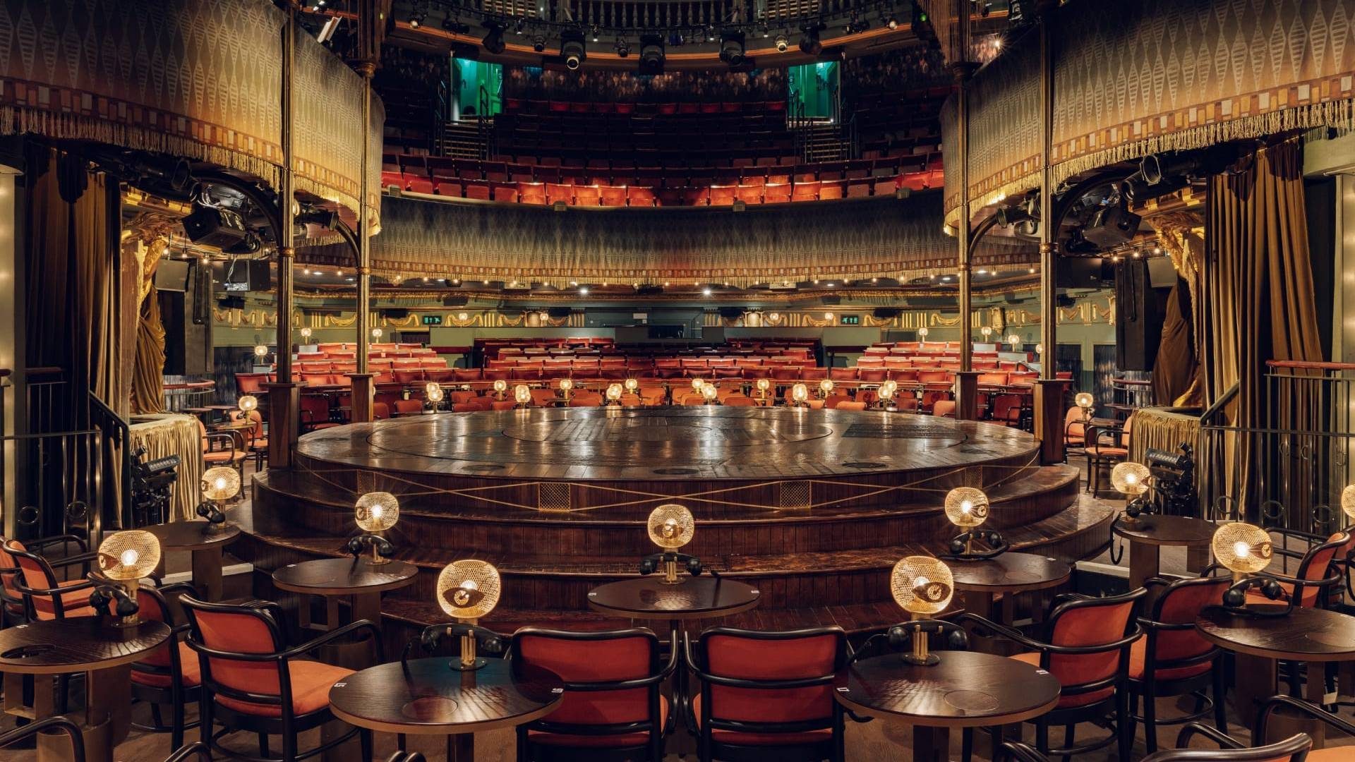 Kit Kat Club. The venue is empty of people. It features a round wooden stage in the centre, surrounded by cabaret tables.